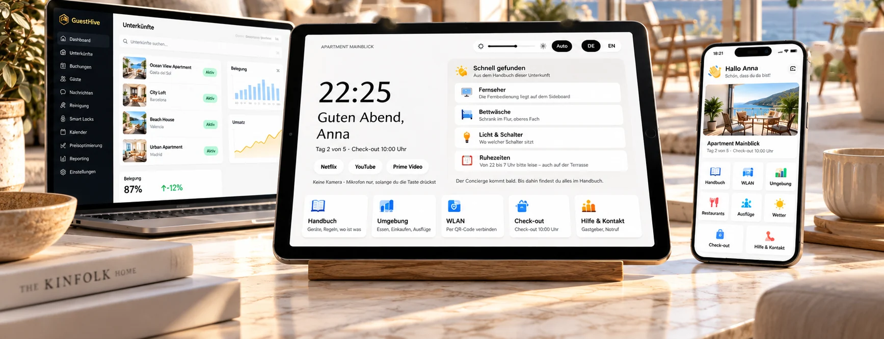 Ein Laptop mit der GuestHive-Verwaltung, ein Tablet auf einem Holzständer mit dem Gästehandbuch von Apartment Mainblick und ein Handy mit der Gäste-App auf einem Marmortisch in einer hellen Ferienwohnung.